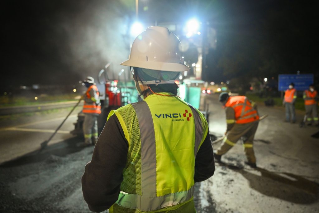 Maintenance worker in yellow high-visibility vest and white helmet looking workers on Via Cristais Highway in background.